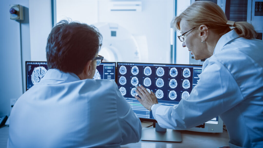 In Control Room Doctor and Radiologist Discuss Diagnosis while Watching Procedure and Monitors Showing Brain Scans Results, In the Background Patient Undergoes MRI or CT Scan Procedure.