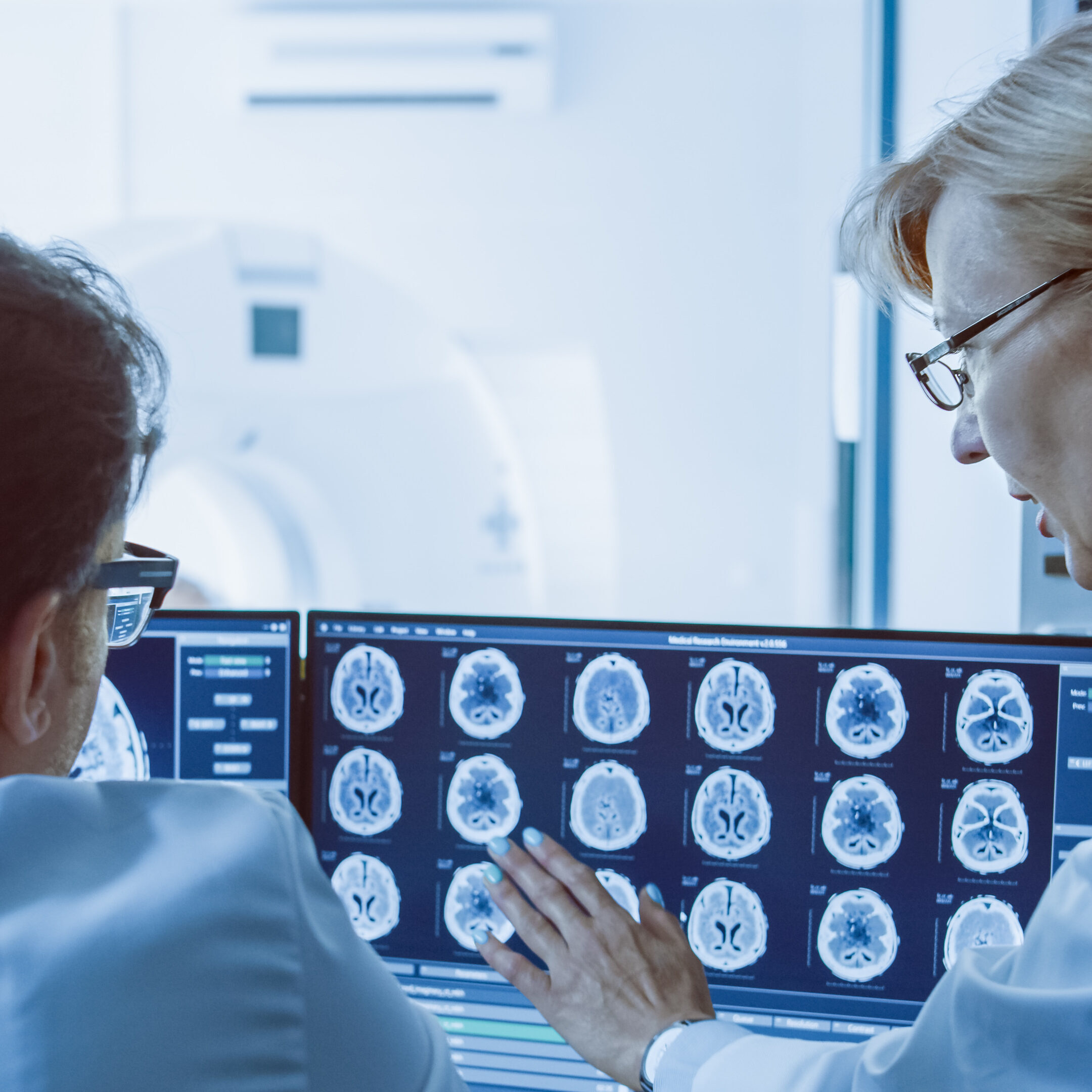 In Control Room Doctor and Radiologist Discuss Diagnosis while Watching Procedure and Monitors Showing Brain Scans Results, In the Background Patient Undergoes MRI or CT Scan Procedure.
