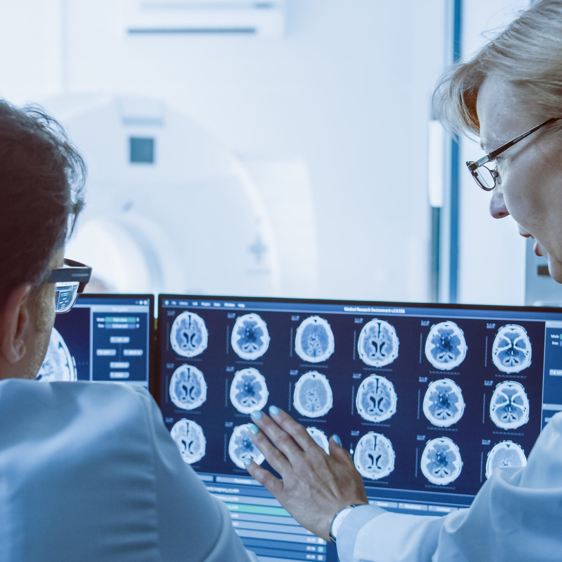 In Control Room Doctor and Radiologist Discuss Diagnosis while Watching Procedure and Monitors Showing Brain Scans Results, In the Background Patient Undergoes MRI or CT Scan Procedure.